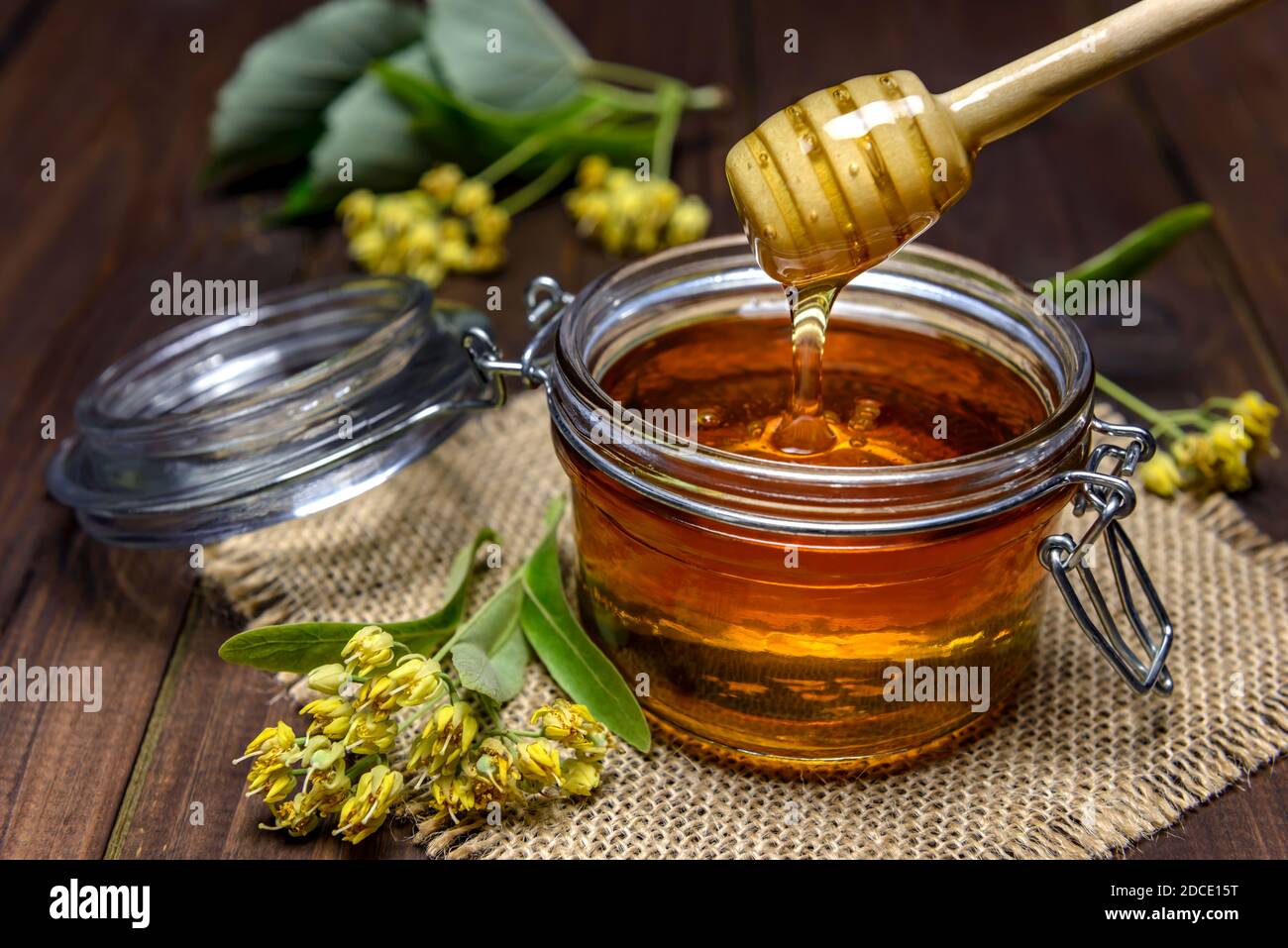 Il miele di Linden sta versando con il bastone di legno in un vaso accanto ai fiori di tiglio. Messa a fuoco selettiva. Foto Stock