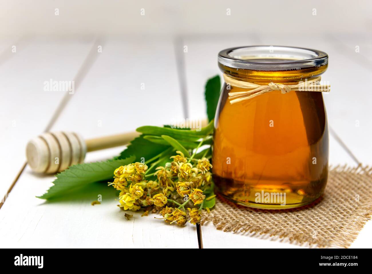 Vaso con miele di tiglio, bastone per miele e fiori di tiglio freschi su un tavolo di legno. Foto Stock