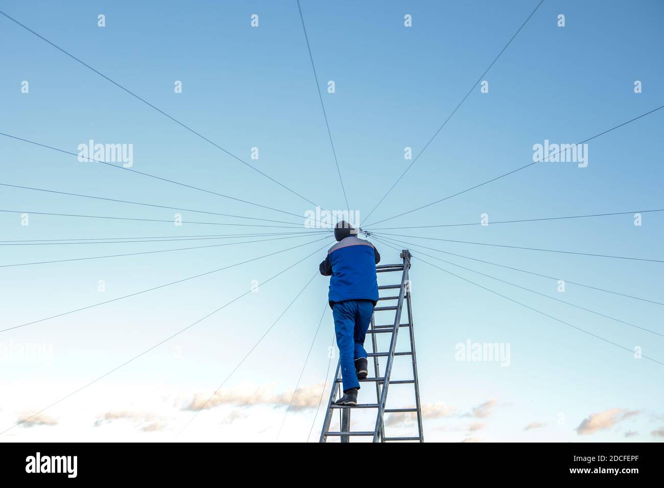 Elettricista ripara il cablaggio elettrico sul tetto di un alto edificio che si erge sulle scale contro il cielo blu. Spazio di copia Foto Stock