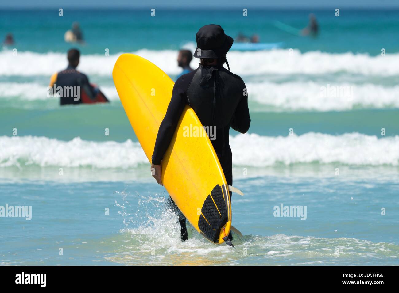 Surfista o uomo non identificabile in una muta che tiene una tavola da surf gialla nelle onde alla spiaggia di Muizenberg, Sud Africa concetto Estate divertimento in vacanza Foto Stock
