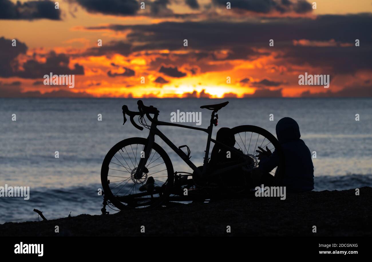 Silhouette di un paio di ciclisti maschili che riposano su una spiaggia mentre si guarda il sole che scende. Persone che guardano il tramonto in mare. Foto Stock