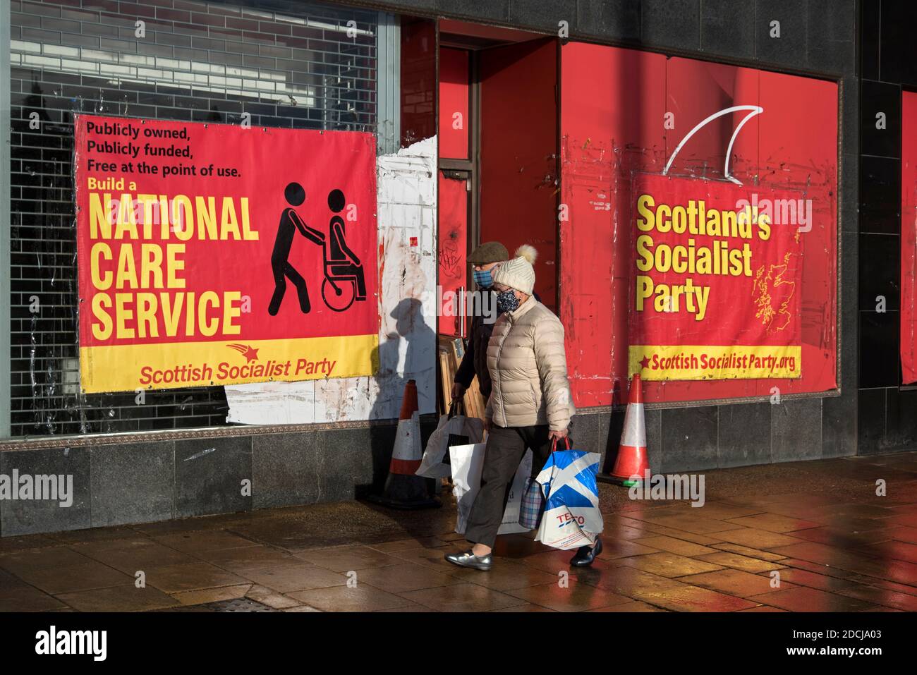 Una coppia anziana che indossa maschere facciali cammina con i manifesti del Partito Socialista Scozzese che chiedono un servizio di assistenza nazionale su Princes Street, Edimburgo. Foto Stock
