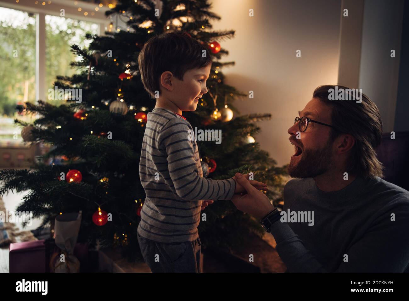 Uomo seduto dall'albero di Natale che si diverte con suo figlio a casa. Padre e figlio si godono a casa durante la vigilia di Natale. Foto Stock