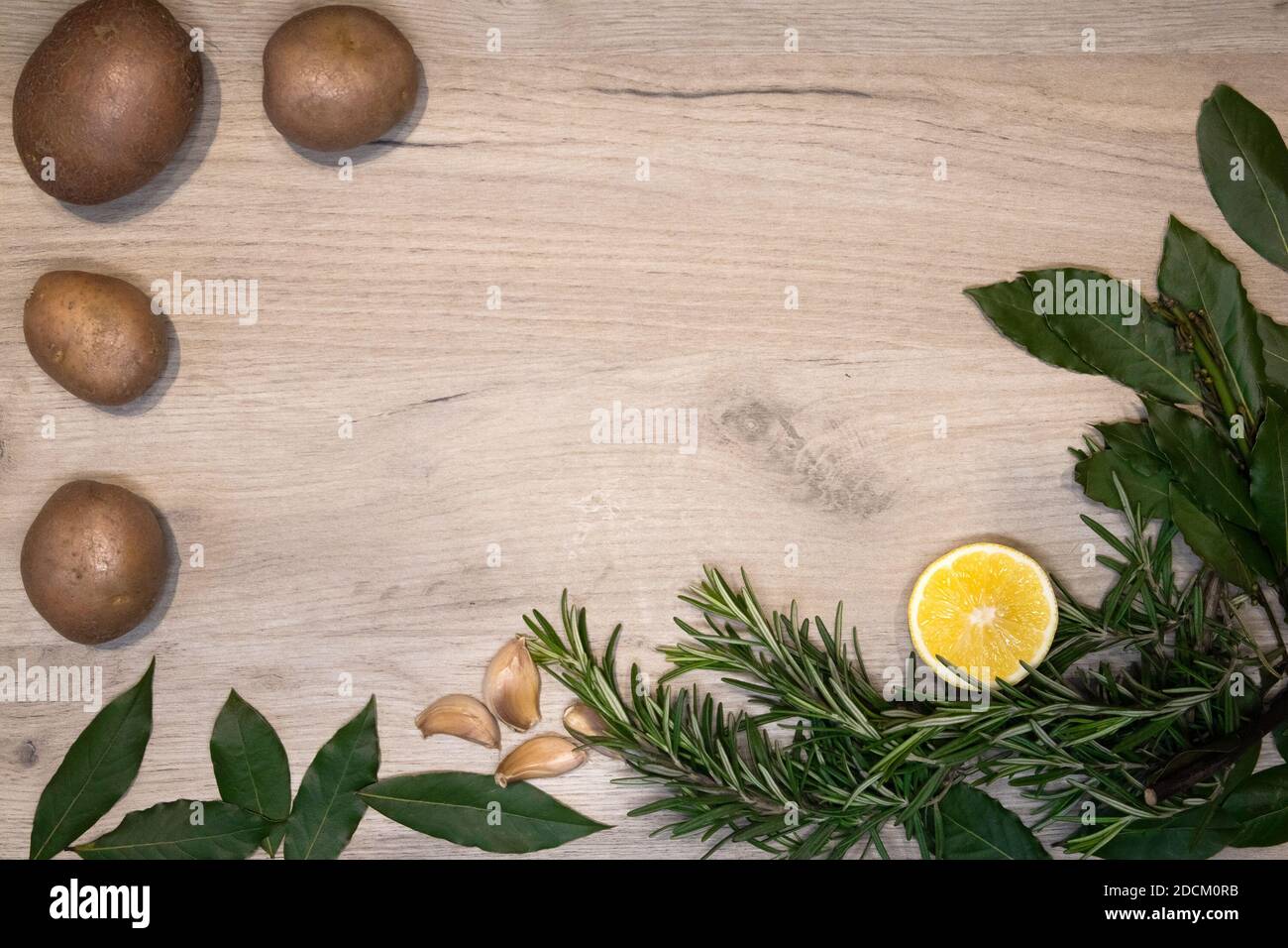 Bordo di legno chiaro incorniciato con sprigs di rosmarino fresco, foglie di alloro, metà di limone, spicchi d'aglio e poche patate crude, modello di disegno Foto Stock