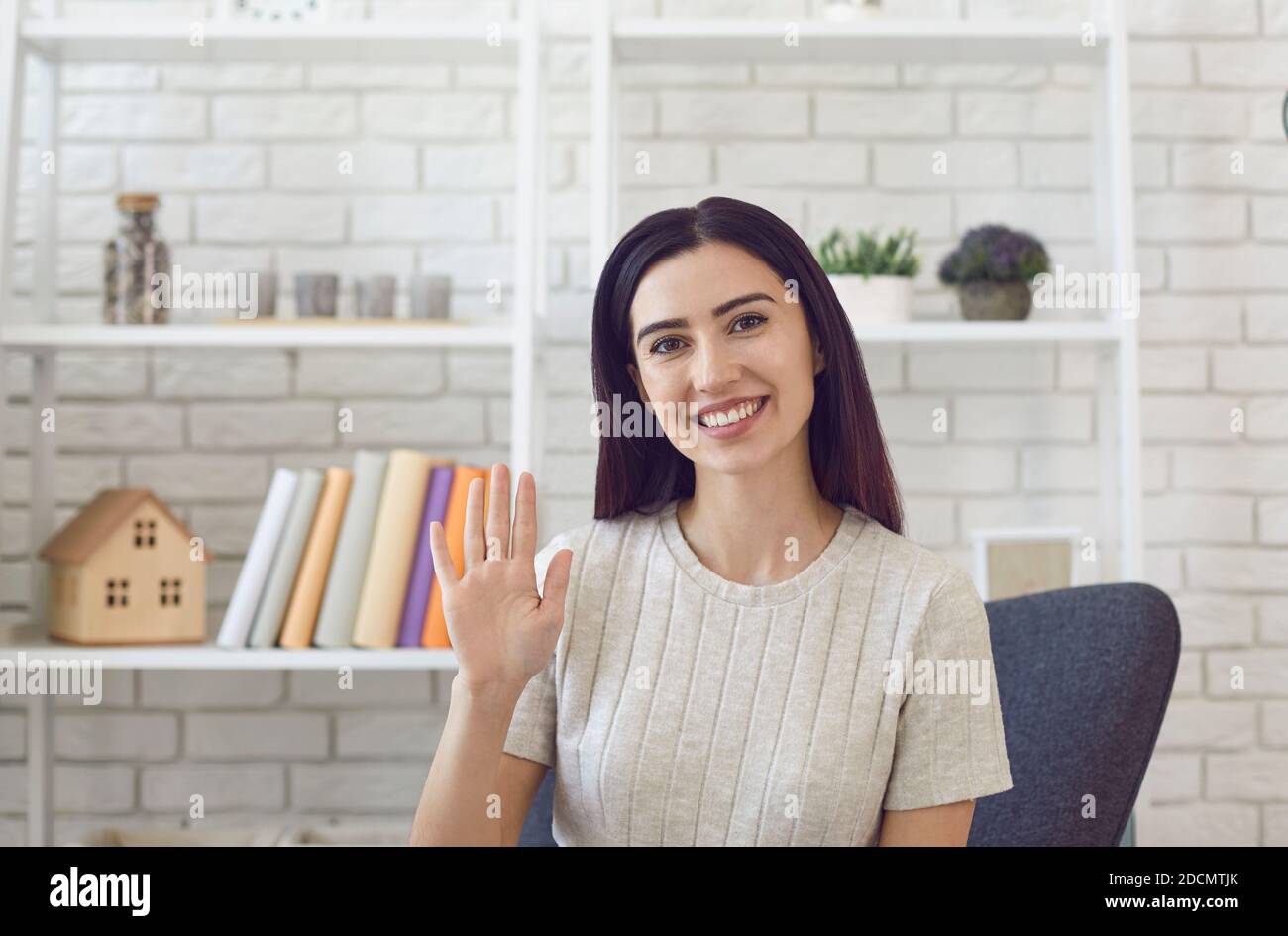 Giovane donna sorridente seduto sulla sedia in un ufficio moderno muovendo la mano verso la fotocamera Foto Stock