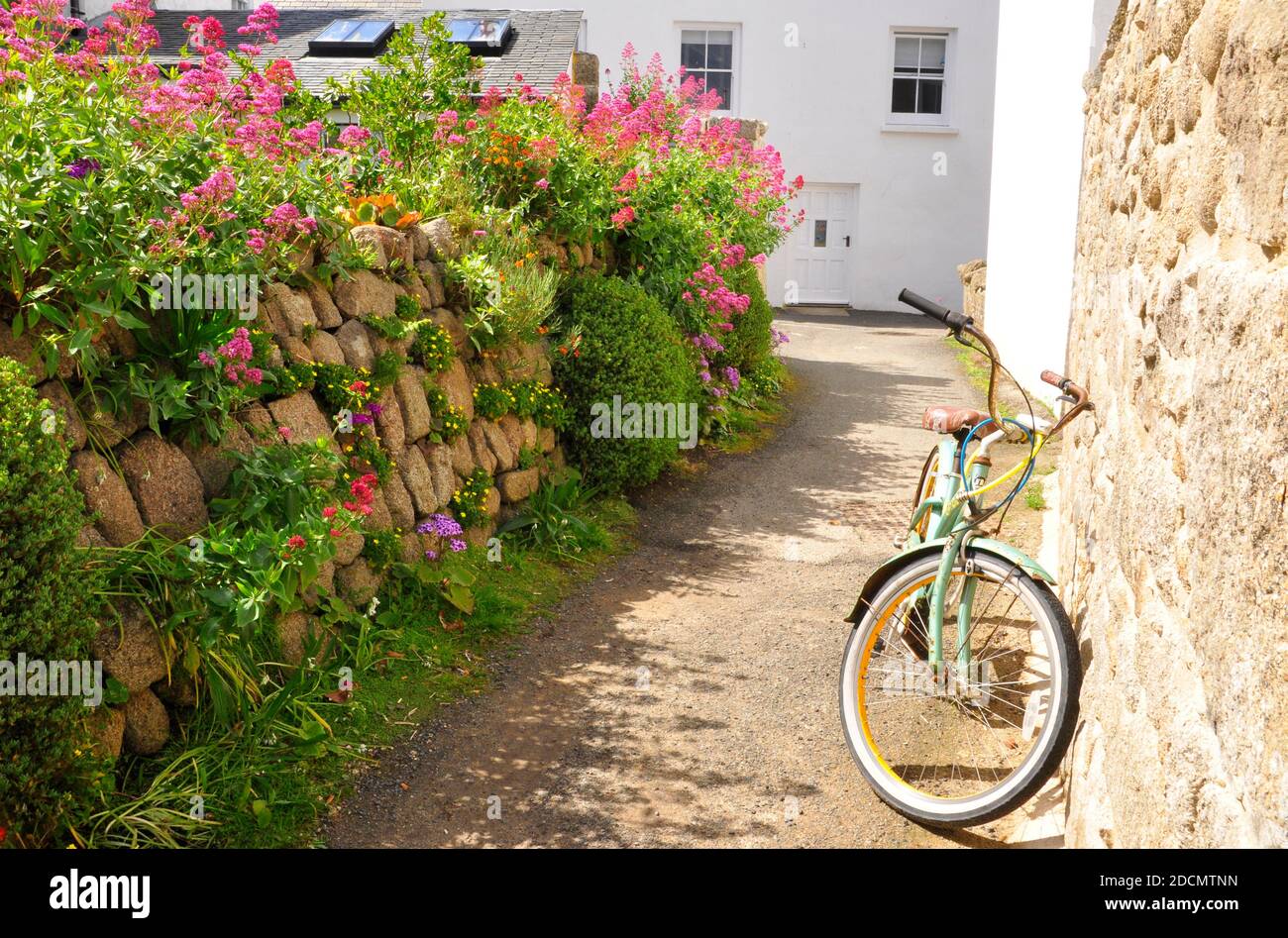 Un tranquillo vicolo posteriore nella città di Hugh su St Mary's, isole di Scilly. Il sole luminoso, la parete di granito ricoperta di fiori di primavera e la bicicletta caratterizzano il Foto Stock