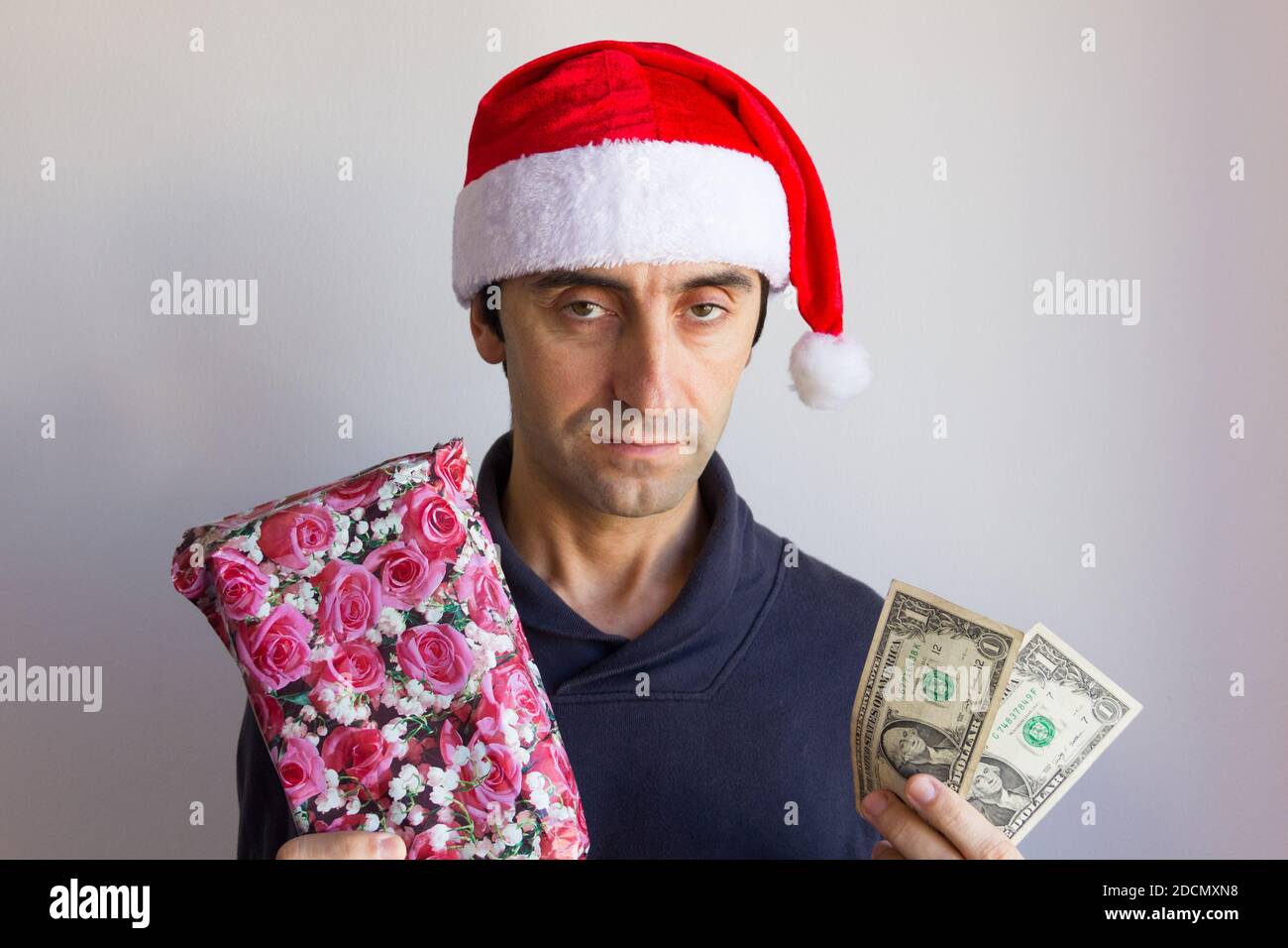 Ragazzo annoiato con il cappello di santa che tiene il regalo avvolto e poco contanti con il volto deluso su sfondo bianco. Natale stagione shopping Foto Stock