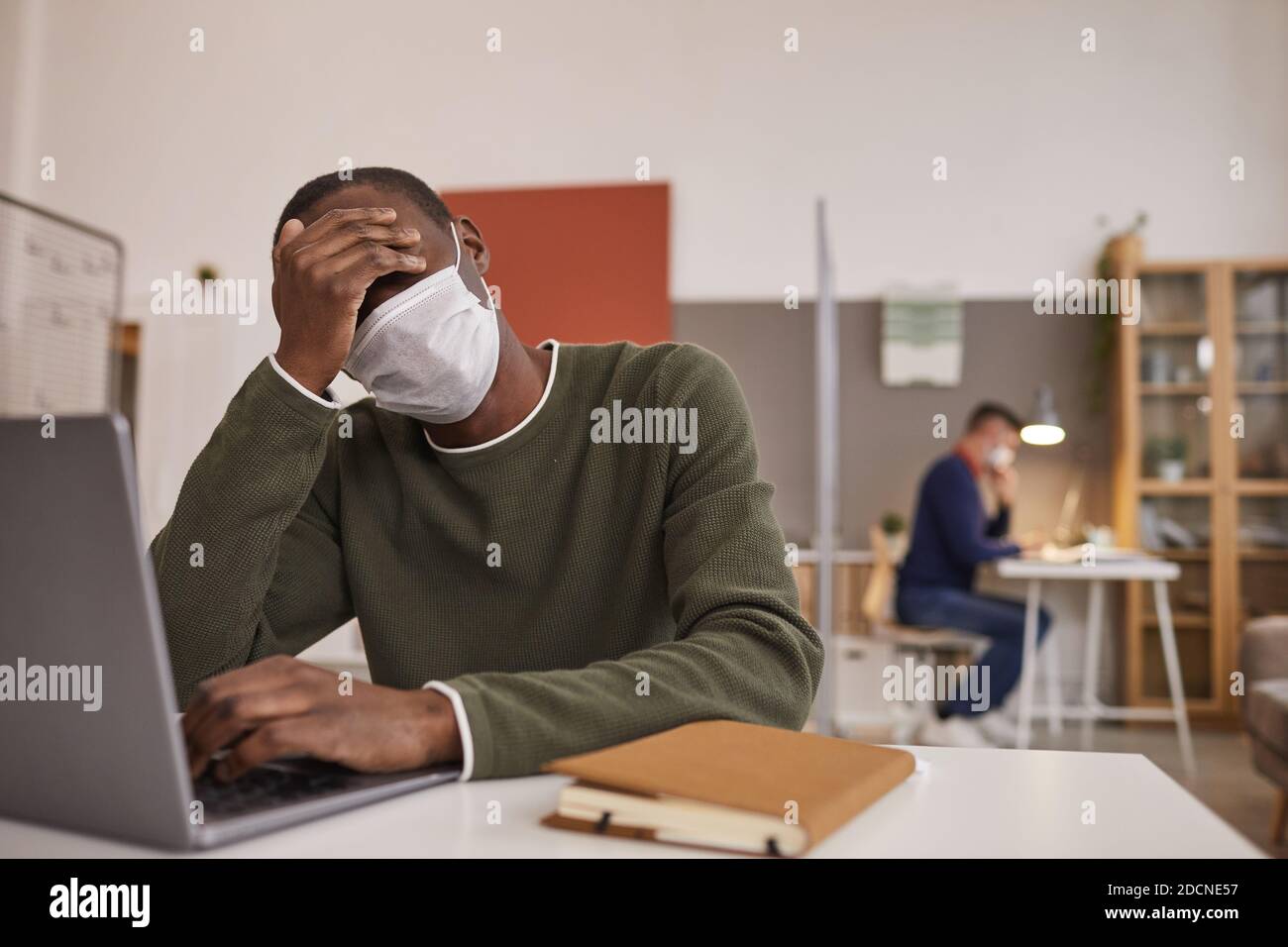 Ritratto di un uomo afro-americano stanco e malato che indossa una maschera e utilizza un computer portatile mentre lavora alla scrivania in ufficio, spazio per la copia Foto Stock