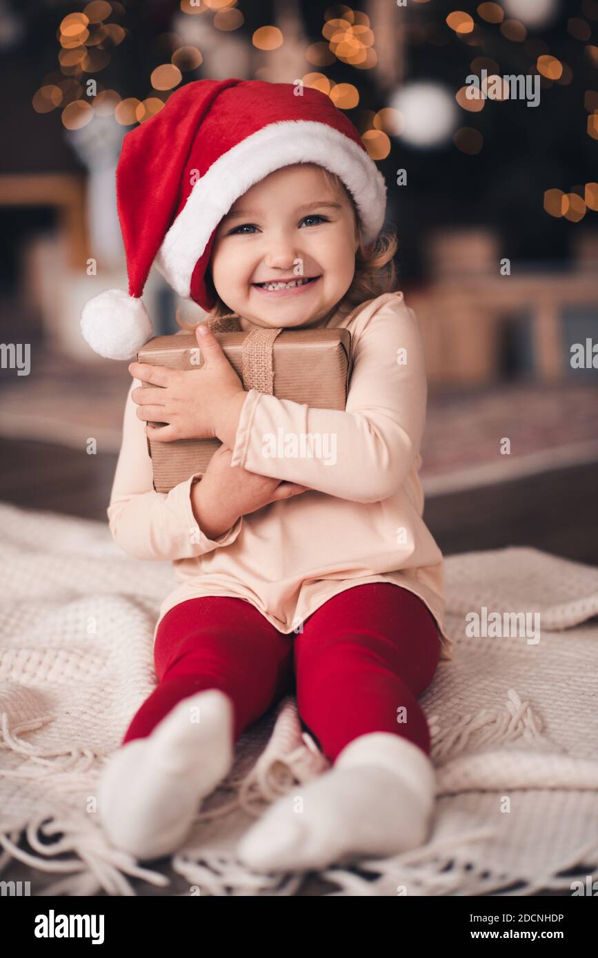 Divertente bambina 3-4 anni tenendo presente scatola di Natale che indossa cappello di babbo natale e vestito seduta sopra l'albero con luci incandescenti primo piano. Cercando un Foto Stock