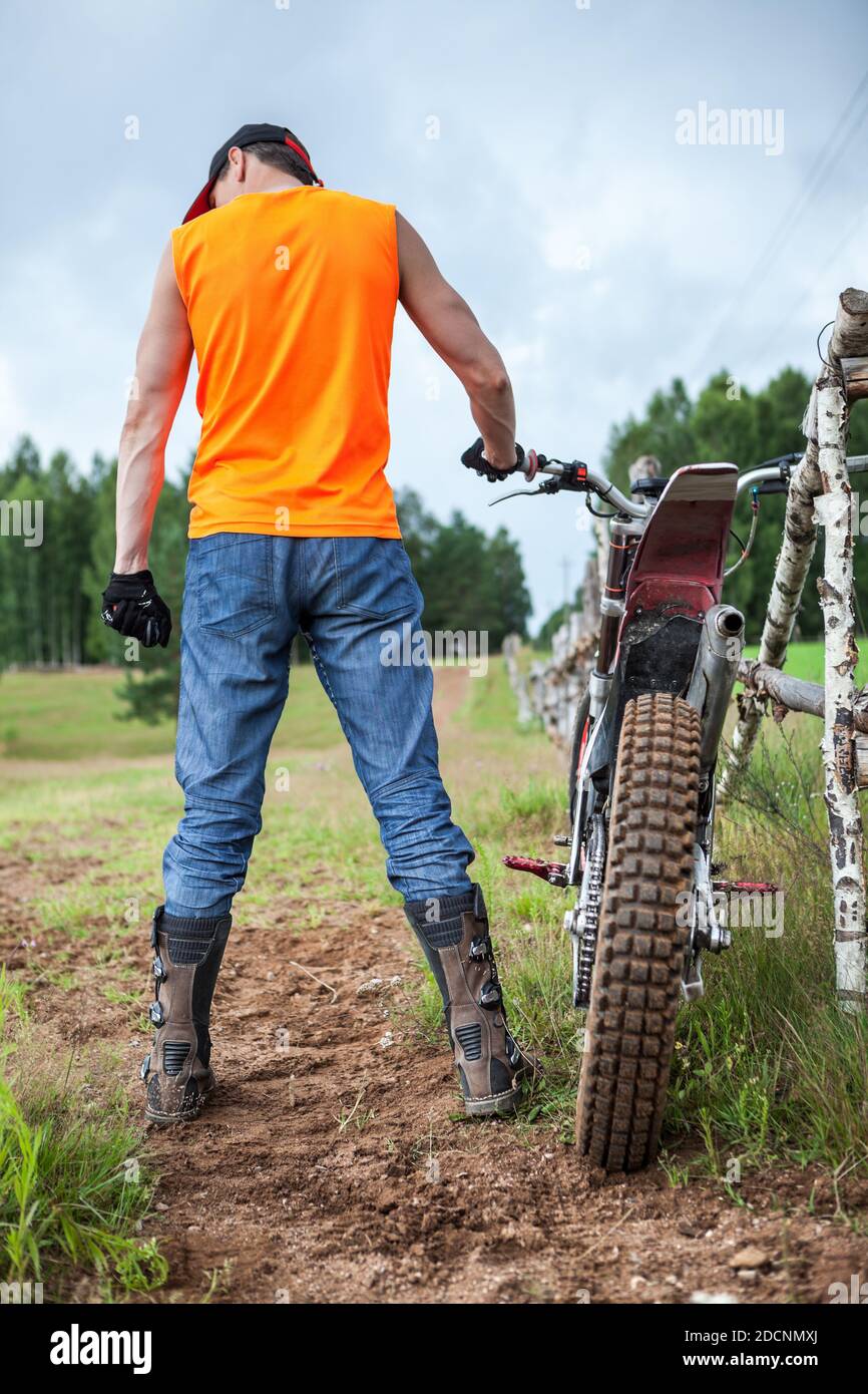 Vista a tutta lunghezza sul retro del pilota irriconoscibile con la sua moto da fondo, la camicia arancione e gli stivali protettivi Foto Stock