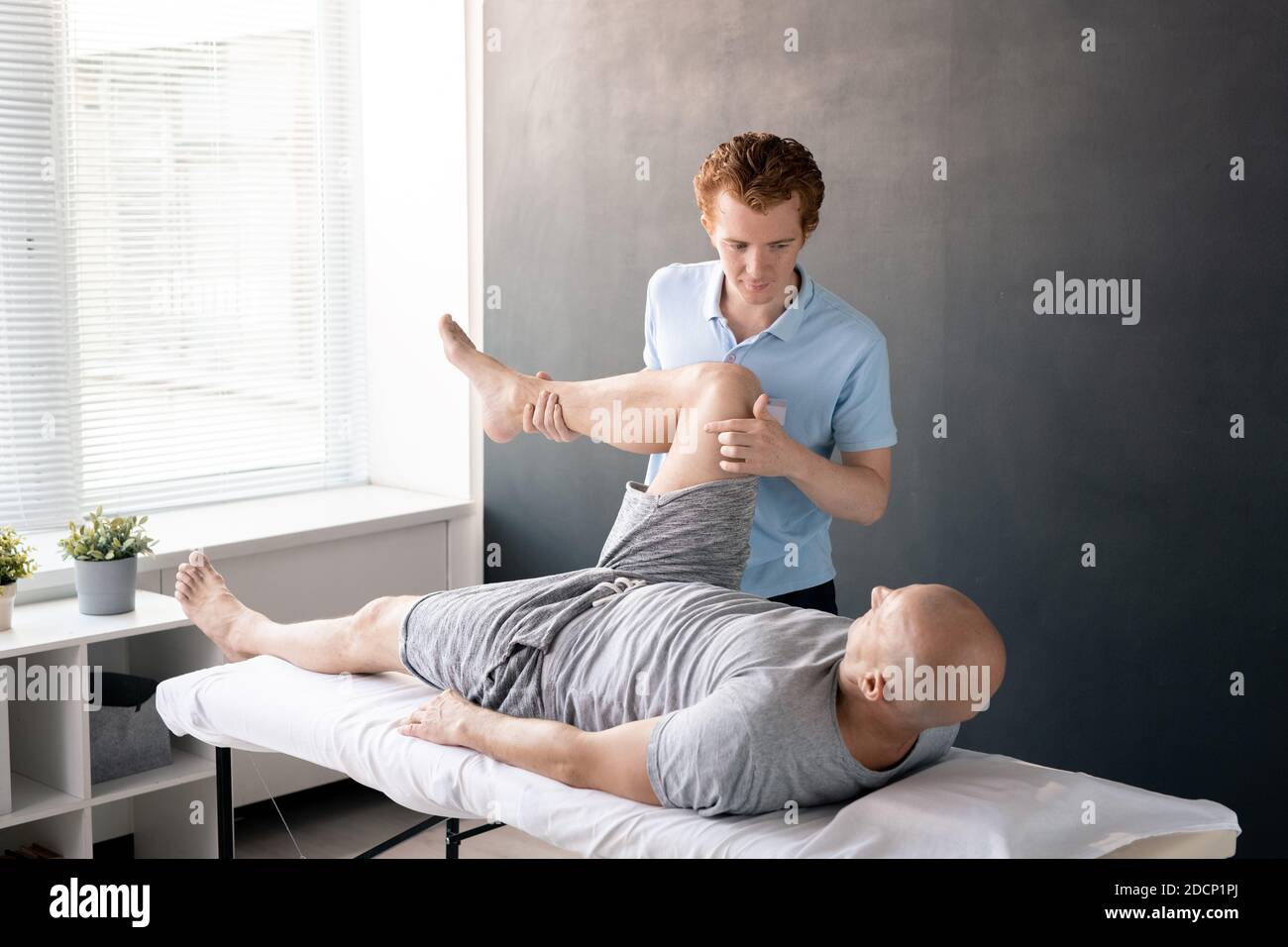 Fisioterapista maschile serio che guarda il paziente con la gamba piegata nel ginocchio Foto Stock