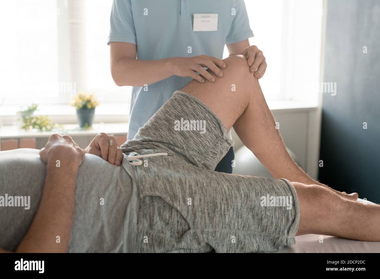 Uomo sdraiato sul divano medico mentre un giovane fisioterapista massaggiava il suo ginocchio sinistro Foto Stock