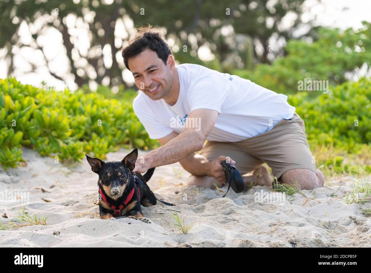 il giovane si inginocchia per accarezzare il cane nero coperto di sabbia in spiaggia Foto Stock