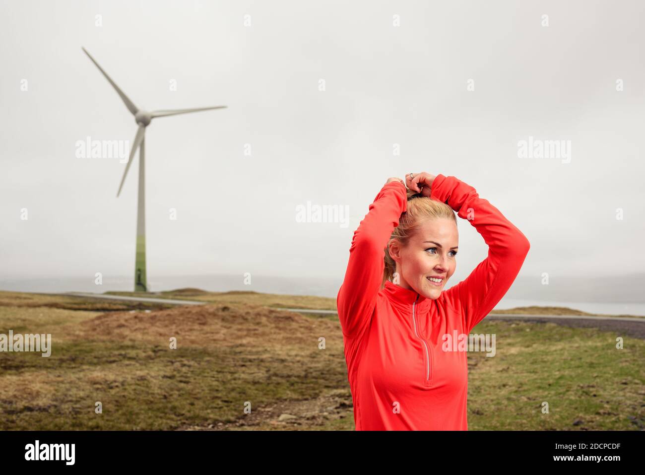 Bionda sportiva sorridente e facendo ponytail vicino turbina eolica durante allenamento Foto Stock