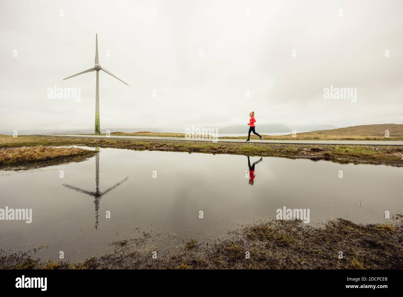Sportswoman che corre sul sentiero vicino al lago e mulino a vento su grigio giorno in natura Foto Stock