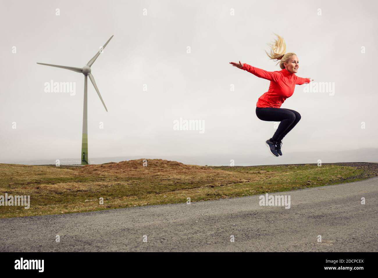 Donna che salta con le braccia allungate durante l'allenamento contro mulino a vento in natura Foto Stock