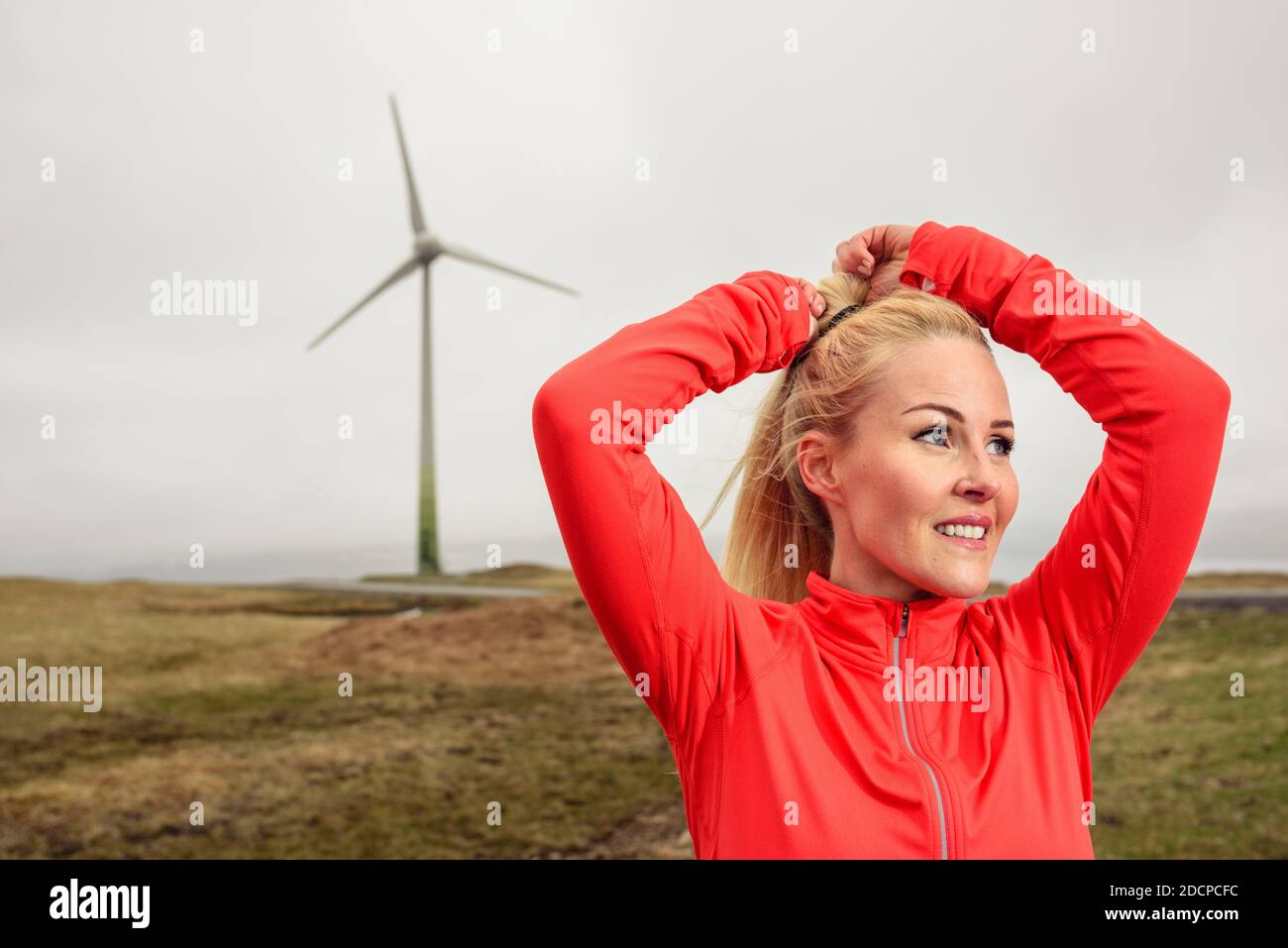 Atleta adulto biondo sorridente e facendo ponytail vicino al mulino a vento dentro campagna Foto Stock