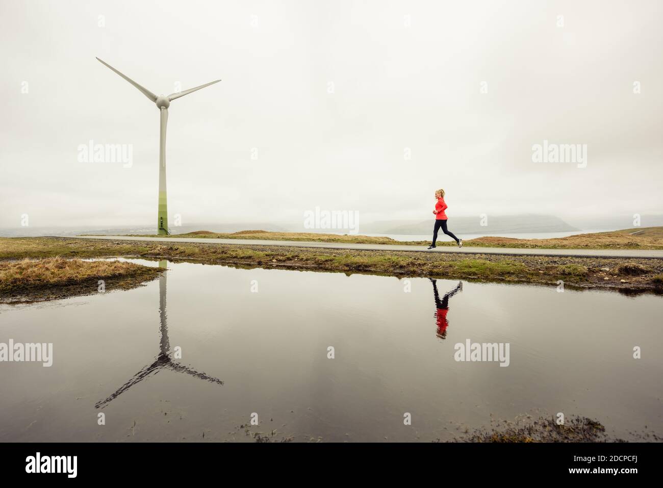 Atleta femmina che fa jogging vicino allo stagno e turbina eolica contro il grigio cielo in natura Foto Stock