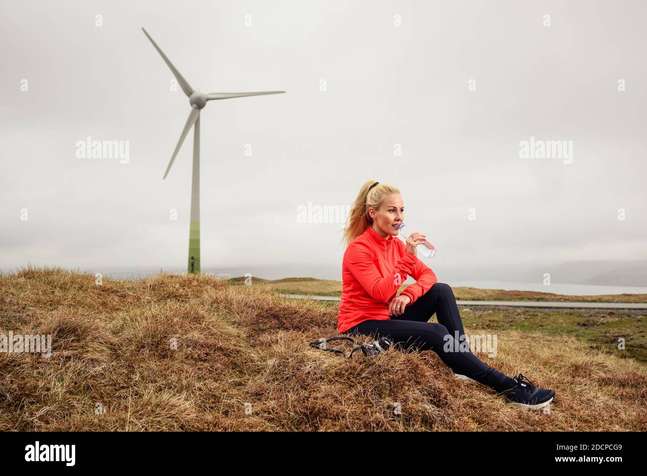 Atleta femmina che sorseggia acqua dalla bottiglia mentre si siede sull'erba vicino al mulino a vento Foto Stock