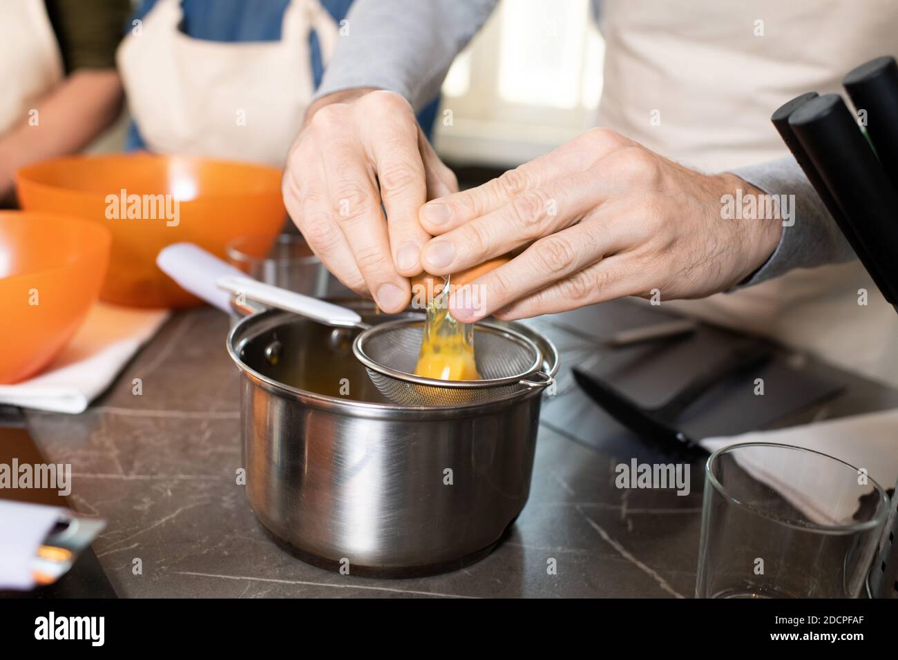 Mani dell'uomo anziano in grembiule che rompono l'uovo nel setaccio sopra il piatto metallico dal tavolo Foto Stock