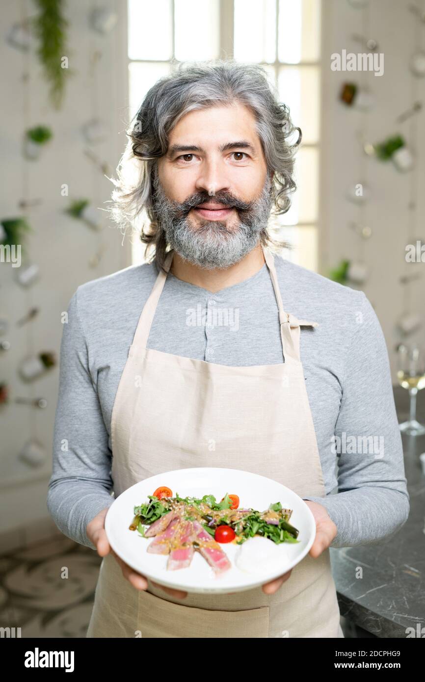 Uomo maturo con la bearded in pullover e grembiule che tiene piatto con insalata di verdure Foto Stock