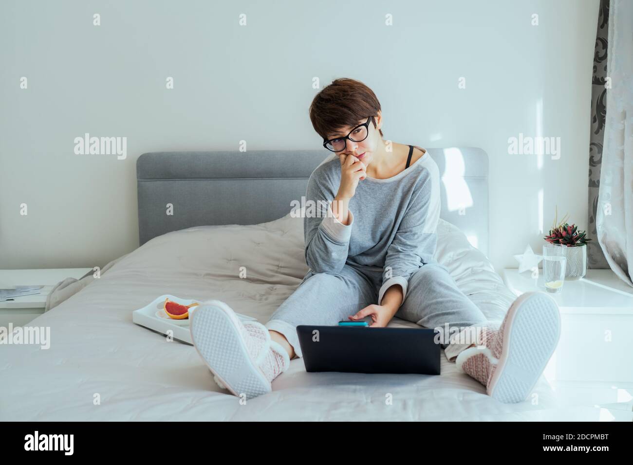 Donna caucasica confusa e premurosa in occhiali e vestiti casual casa utilizzando un computer portatile seduto sul suo letto e facendo colazione. Funzionamento remoto da Foto Stock