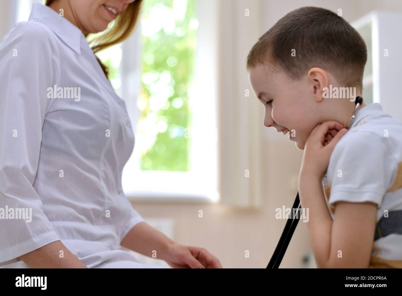 Il bambino ride con le cuffie stetoscopiche nelle orecchie, insieme al medico una ragazza in abiti bianchi. Foto Stock