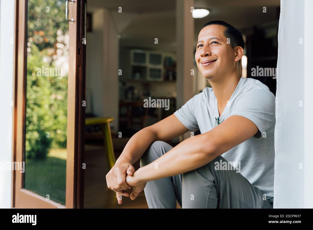 Ritratto di 30-40 anni felice sorridente e pensieroso uomo seduto sulla porta d'ingresso a casa in estate. Razza mista di ragazzo asiatico e caucasico che guarda via - con spazio di copia Foto Stock