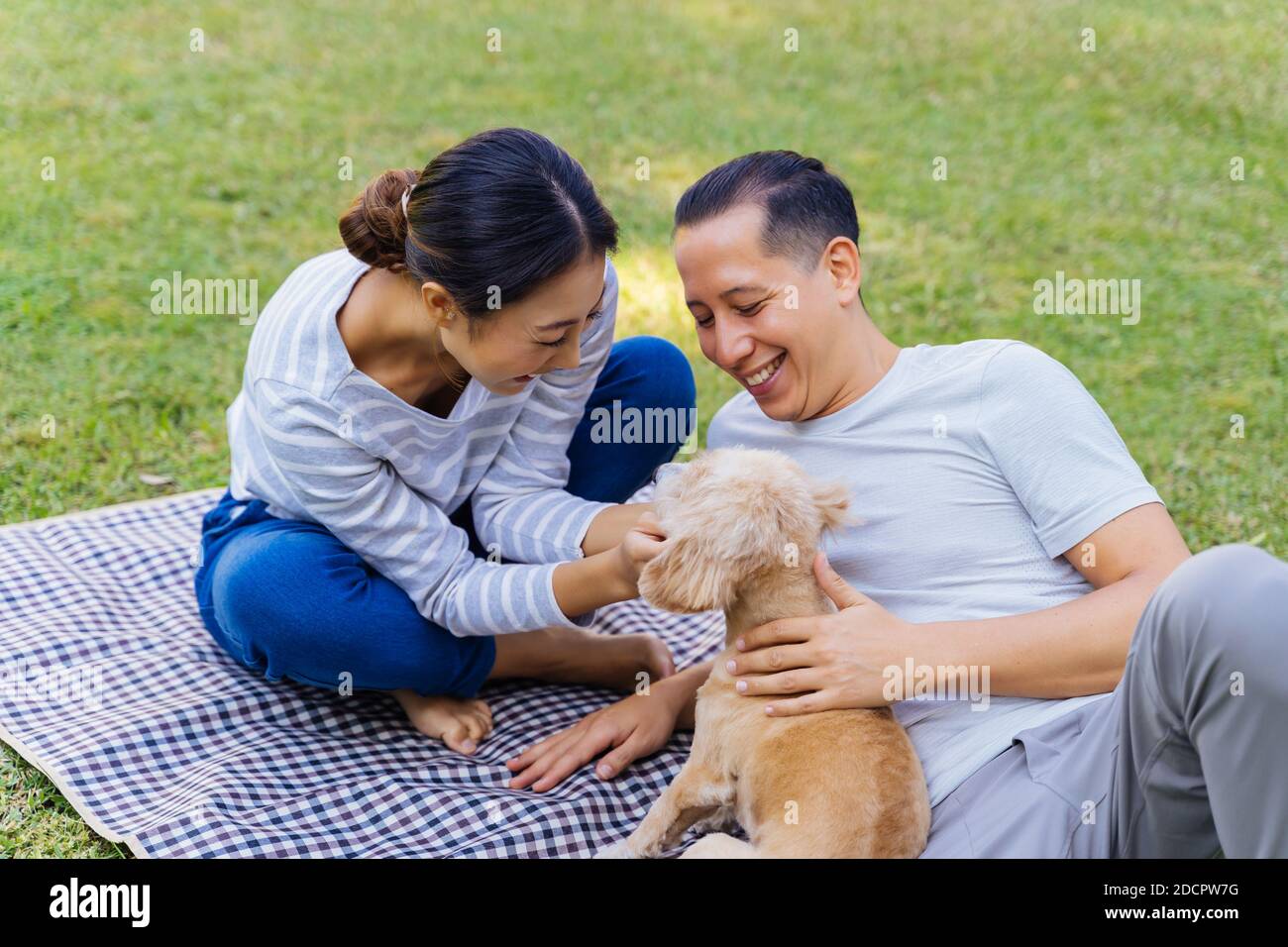 Giovane coppia asiatica adulta che gioca un cucciolo con erba verde all'aperto in background. anni 30, uomo e donna maturi che si stendono sul tappeto nel parco toccando un cane animale domestico. Famiglia felice e concetto di vita moderna Foto Stock