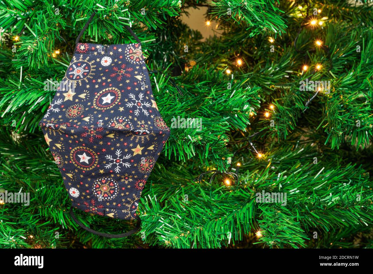Natale covid-19 maschera appesa come decorazione su albero verde con luci. Il disegno di protezione del viso del coronavirus come ornamento stagionale all'albero artificiale. Foto Stock