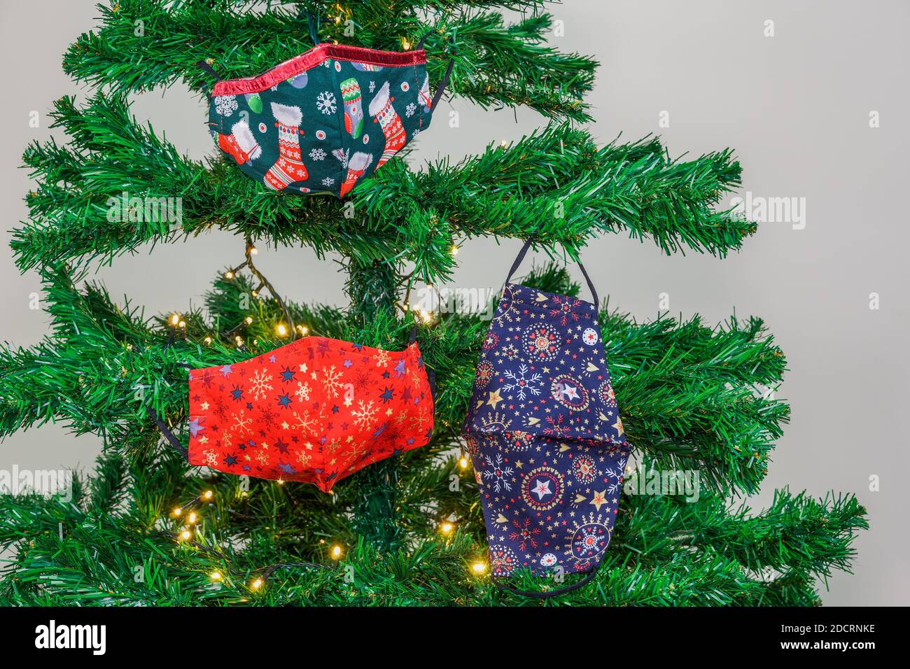 Natale covid-19 maschere appendere come decorazione su albero con luci. La protezione del viso del coronavirus festivo si disegna come ornamenti stagionali sull'albero artificiale. Foto Stock