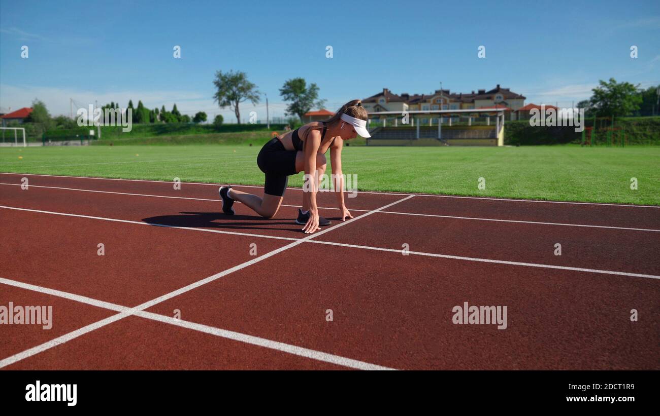 Giovane FIT femmina sprinter inizio corsa su pista a stadio in sole giornate estive. Vista laterale della donna atletica attiva che pratica la posizione sprinter preparazione per la corsa. Concetto di sport. Foto Stock