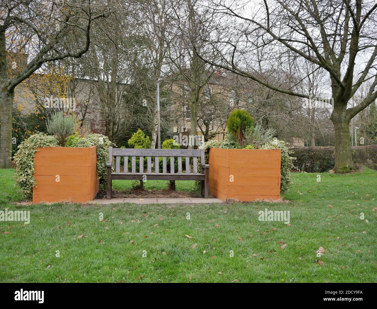Panca di parco di legno con ricordo scolpito in traversa con grandi piantatrici marroni su ogni lato con arbusti d'erba intorno agli alberi sullo sfondo Foto Stock