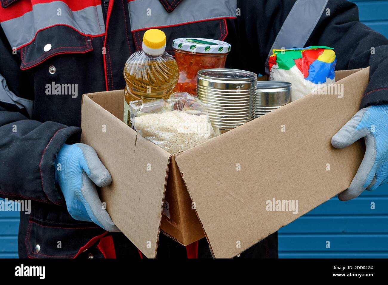 Un volontario in una giacca invernale contiene una scatola di cartone con cereali secchi, cibo in scatola, burro e altri prodotti. Aiuto durante una pandemia. Donazioni di fo Foto Stock
