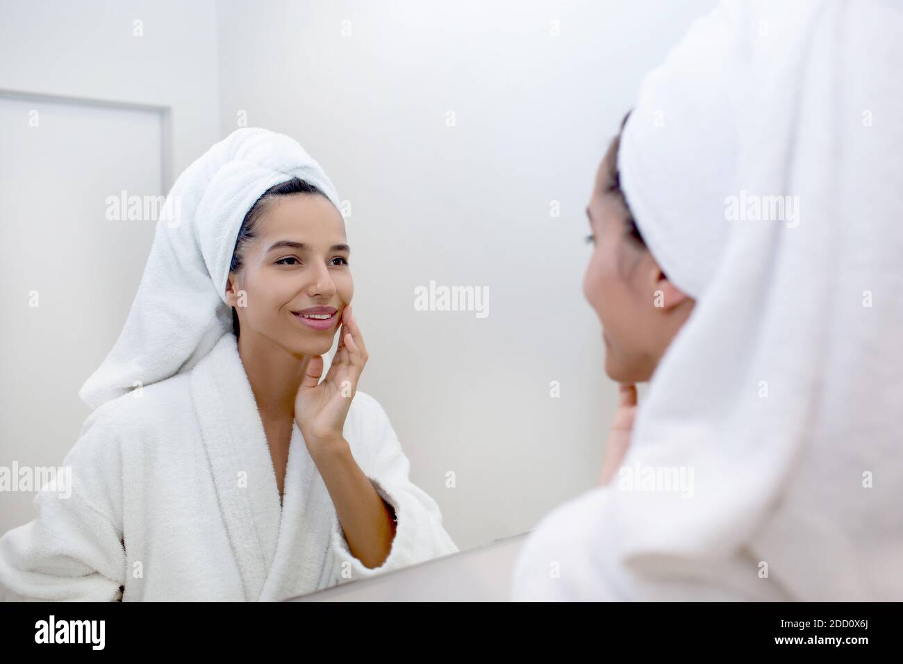 Ritratto di bella donna di razza mista riflessa nello specchio indossando accappatoio toccando la pelle pulita e fresca del viso dopo il lavaggio Foto Stock