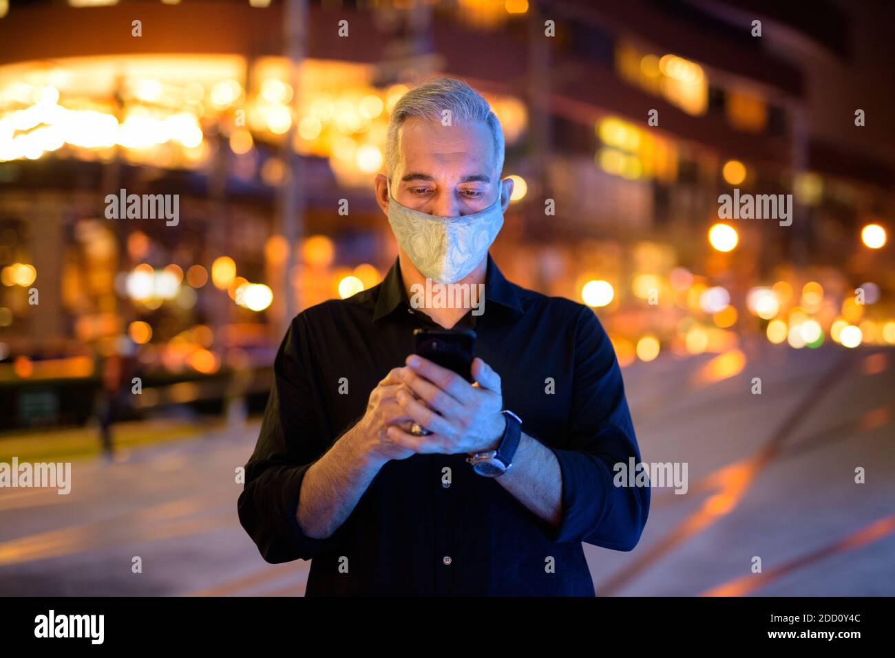 Uomo di notte per strada con maschera facciale protettiva per proteggere dal virus della corona di covid 19 mentre si usa il telefono Foto Stock