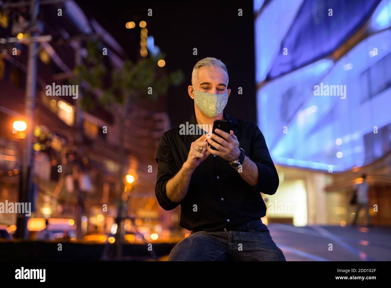 Uomo di notte per strada con maschera facciale protettiva per proteggere dal virus della corona di covid 19 mentre si usa il cellulare telefono Foto Stock