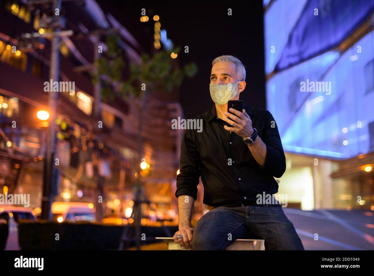 Uomo di notte per strada con maschera facciale protettiva per proteggere dal virus della corona di covid 19 mentre si usa il cellulare telefono e pensiero Foto Stock
