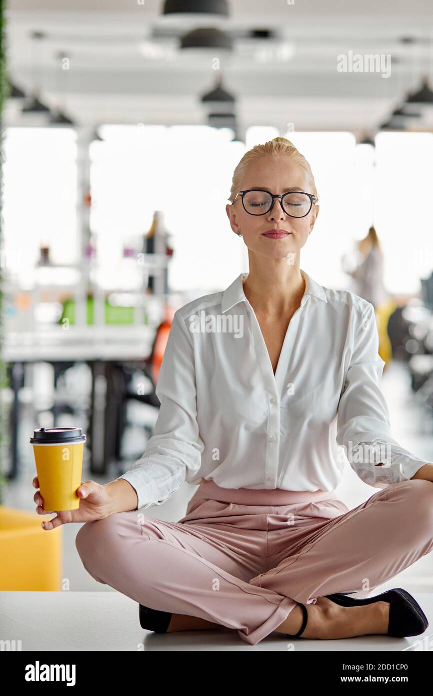 donna in loto posa sul tavolo al posto di lavoro, prendere una pausa, tenendo in mano una tazza di caffè, meditando e rilassandosi Foto Stock