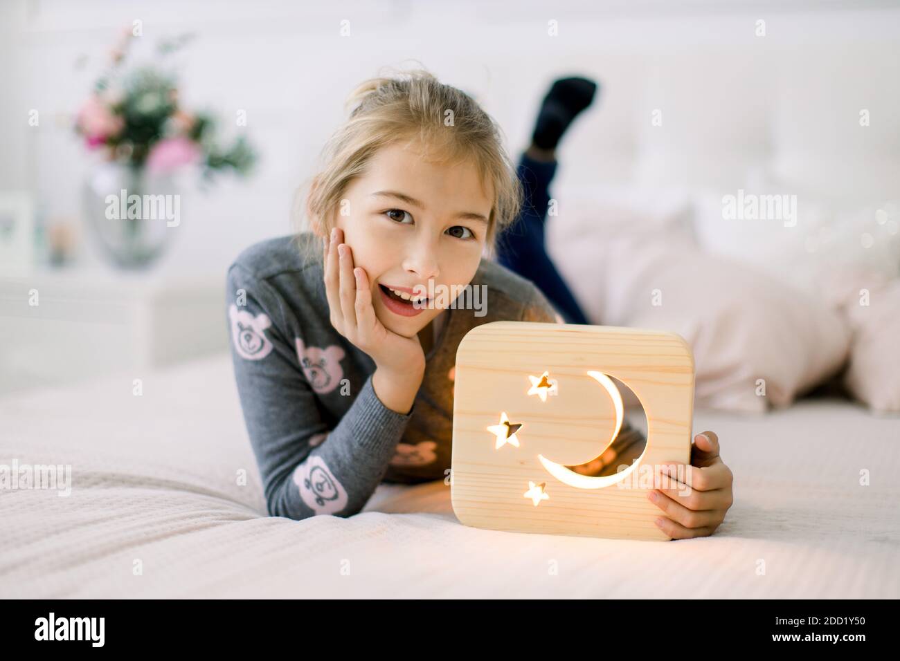 Piccola bella ragazza di 10 anni, sdraiata sul letto in una camera da letto  accogliente luce a casa, e in posa a macchina fotografica con elegante  lampada da notte in legno con