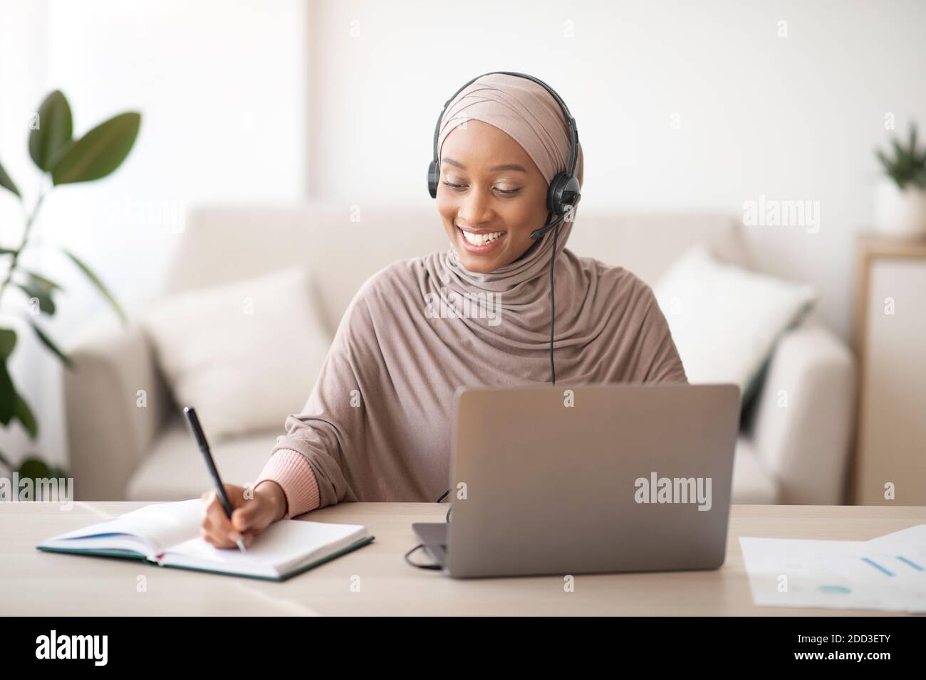 Donna musulmana nera in hijab e cuffia con videoconferenza sul portatile, prendendo appunti durante la lezione online a casa Foto Stock