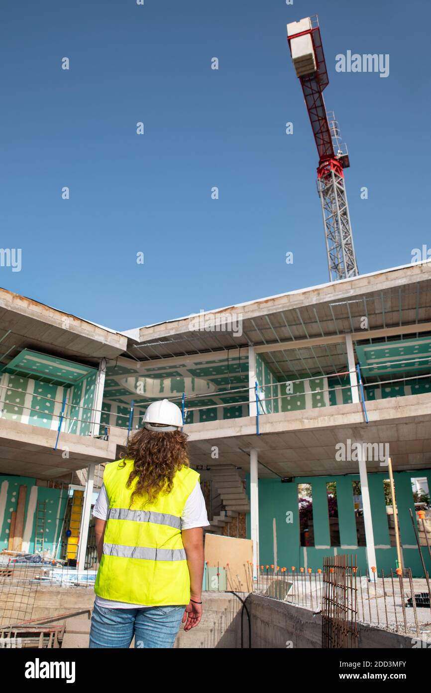 Vista posteriore di una giovane donna architetto con un neon giacca di sicurezza e un elmetto bianco mentre si guarda verso un cantiere Foto Stock