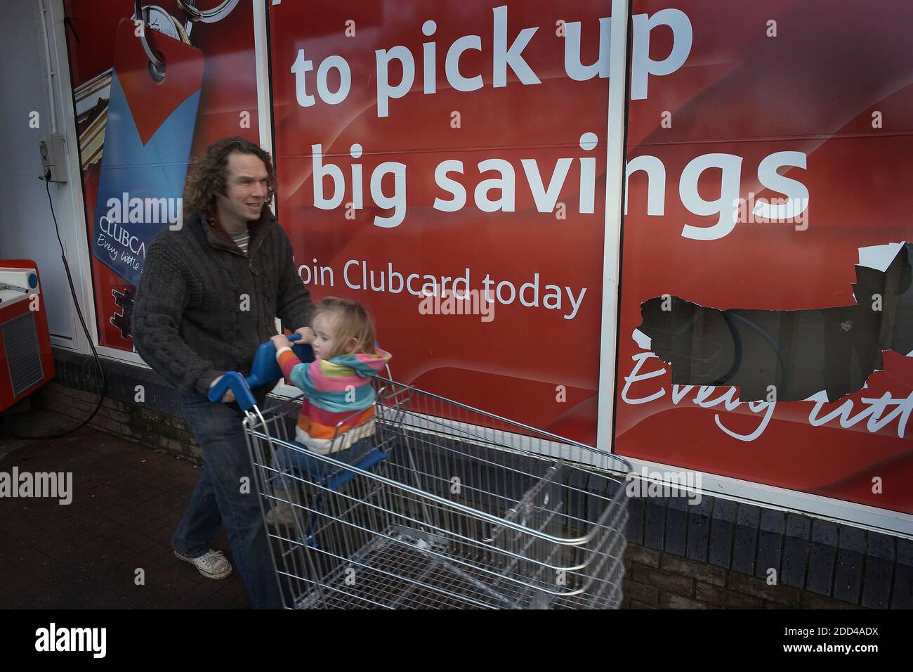 GRAN BRETAGNA / Inghilterra/Bristol.UN uomo con un bambino seduto in un carrello per lo shopping cammina oltre un cartello del supermercato Tesco. Foto Stock