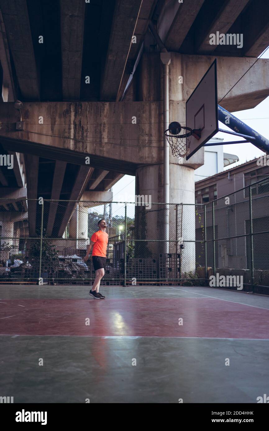 Giovane caucasico che gioca a basket da solo su pista urbana sotto il ponte dell'autostrada indossa una t-shirt arancione Foto Stock