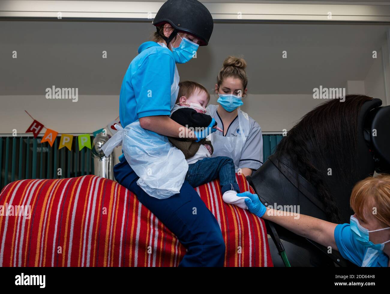 East Lothian, Scozia, UK, NHS Lothian & Muirfield Riding Therapy festeggia 10 anni di ippoterapia: Il fisioterapista Heather Falconer ha aperto la strada al Children’s Therapeutic Riding Service, che fornisce gratuitamente un'equitazione terapeutica a bambini, giovani e adulti con disabilità. Il servizio è stato il primo in Scozia per i bambini con problemi di mobilità, equilibrio, tono muscolare e coordinazione. Oreo, un cavallo meccanico, permette ai giovani di divertirsi con la fisioterapia. Jamie Burns, di 2 anni, cavalca il simulatore meccanico di cavalli Racewood con Lizzie, fisioterapista Foto Stock