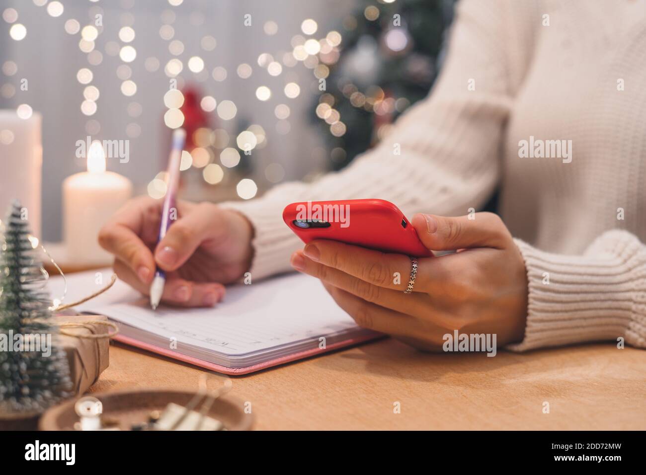 Obiettivi piani fare a fare e lista dei desideri per il nuovo anno natale concetto di scrittura in notebook. Donna mano che tiene la penna sul blocco note e cellulare a casa su wi Foto Stock