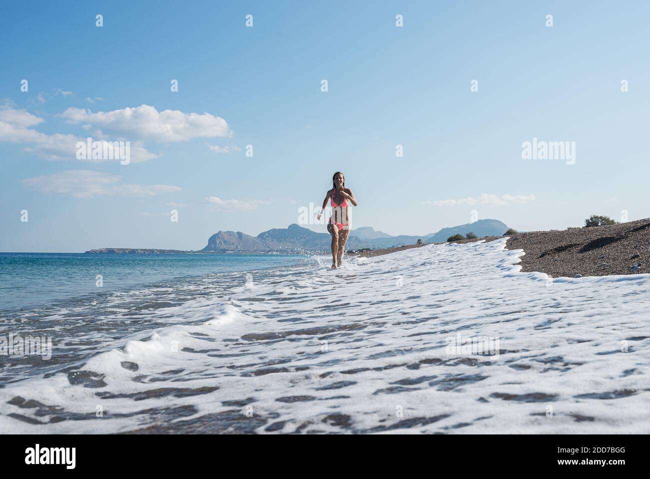Giovane donna in bikini rosa che corre lungo una bella spiaggia di ghiaia con onde schiumose che entrano. Foto Stock