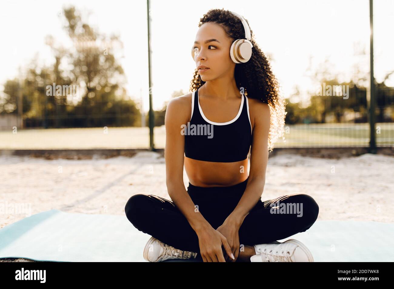 Giovane donna sportiva che ascolta la musica con le cuffie, che fa yoga in posizione lotus, sul tappetino fitness all'aperto nel parco Foto Stock