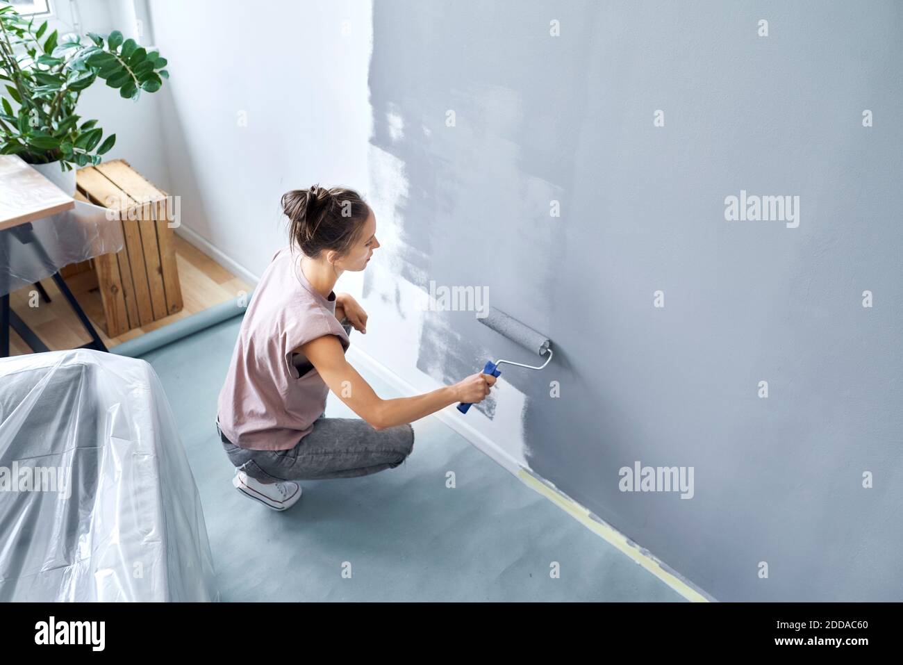 Giovane donna con vernice rullo pittura muro mentre accovacciando a. casa Foto Stock