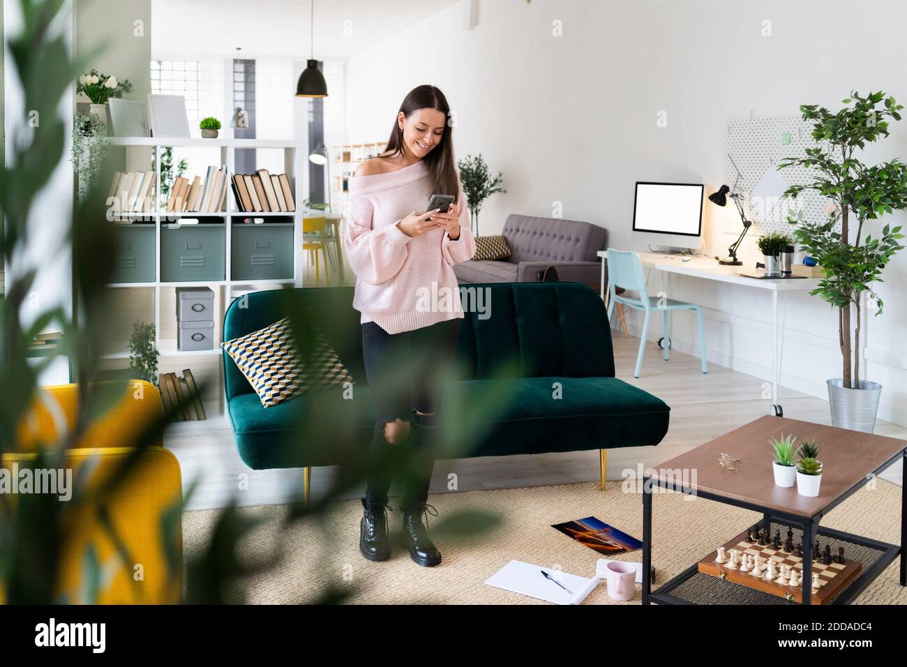 Messaggi di testo sorridenti per le donne tramite smartphone mentre si è in piedi soggiorno a casa Foto Stock